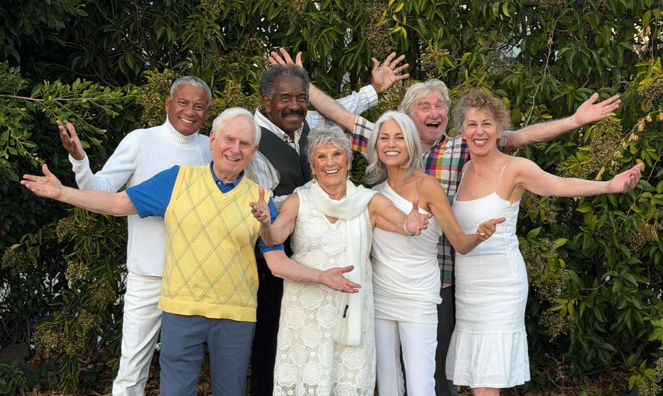 Group of people with arms outstretched, smiling in front of green foliage.
