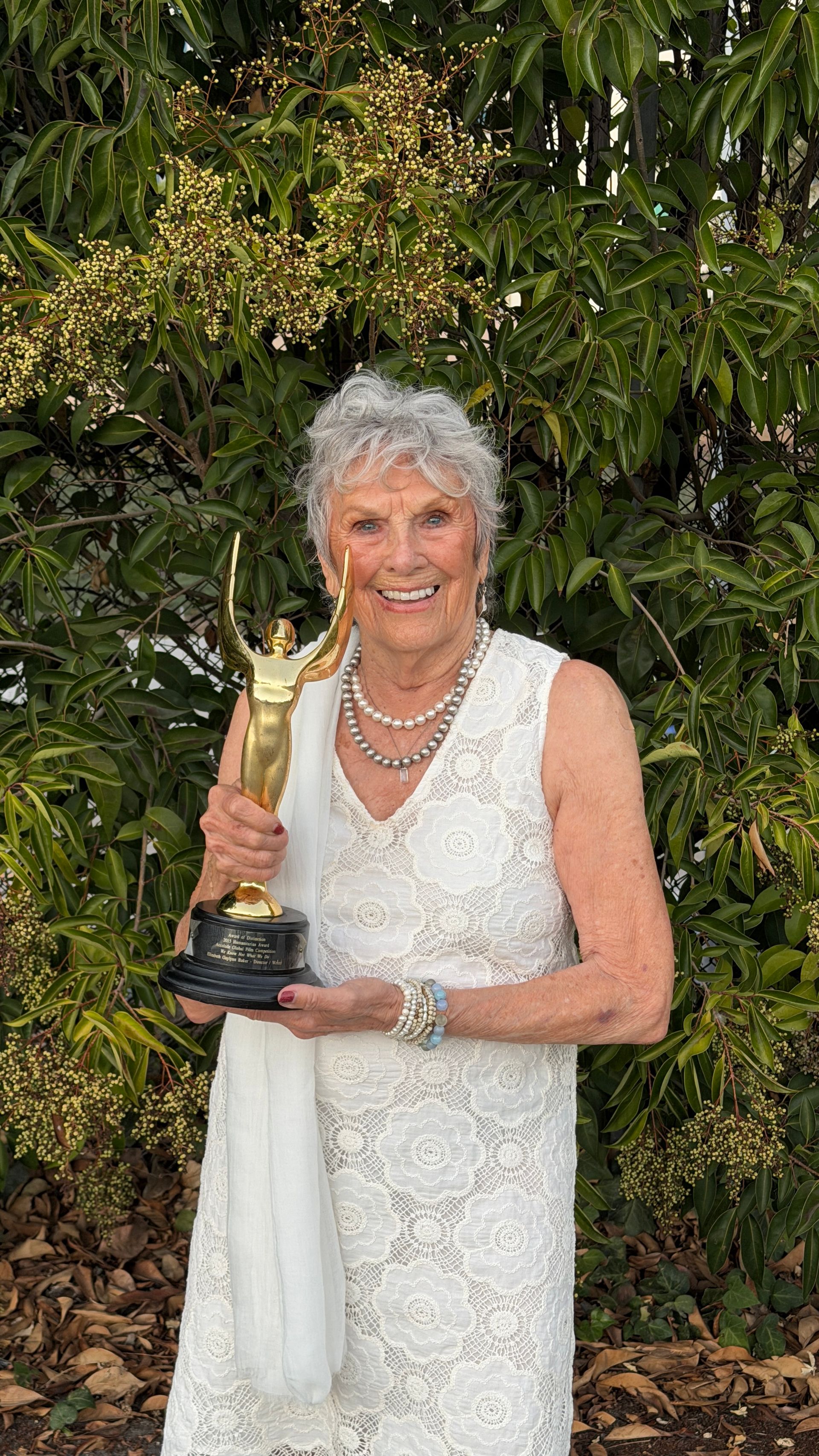 Woman in white dress holds a gold trophy, smiles outdoors in front of leafy green foliage.